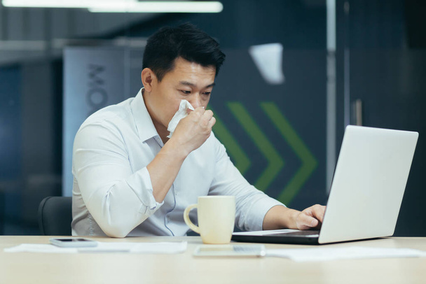 A young Asian freelancer, an office worker fell ill at work. He is sitting at the table, has a runny nose, wipes his nose with a napkin, feels bad, drinks hot tea, medicine. - Photo, image