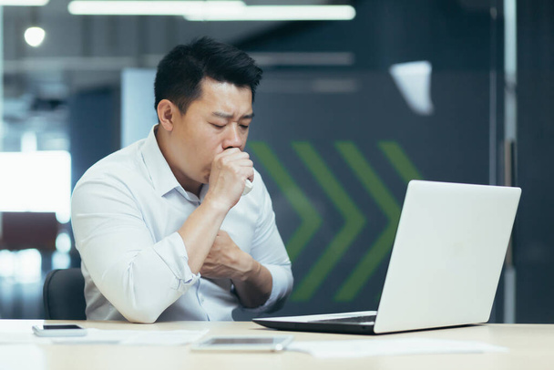A young Asian freelancer, an office worker fell ill at work. He sits at the table and coughs, covers his mouth with his hand, holds his chest, feels bad. - Photo, image