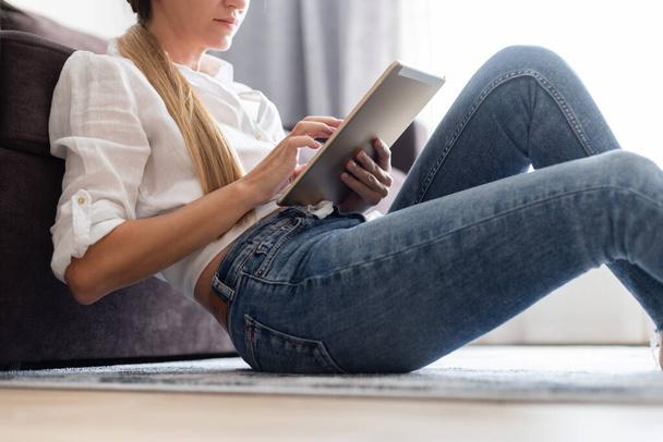 Женщина в белой блузке и джинсах сидит дома на полу и смотрит что-то на планшете. Использование технологии. - Фото, изображение