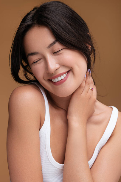 Cheerful young Asian female smiling with closed eyes and touching neck after hearing flattering compliment against brown background - Foto, imagen
