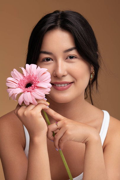 Glad Asian woman with black hair touching face with fresh gerbera flower and looking at camera with smile against brown background - Foto, immagini