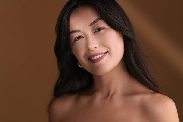 Happy young Asian female with bare shoulders smiling and looking at camera while standing in sunbeam against brown background - Foto, imagen