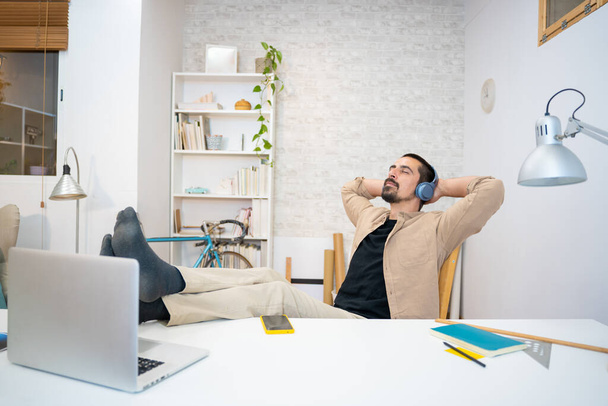 Positive young man keeping hands behind head while listening to music in headphones at home. resting after work in his studio. Creative designer thinking in new ideas. High quality photo - Photo, Image