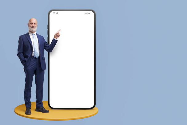 Confident businessman pointing at a big blank smartphone screen, business and online financial services concept - 写真・画像
