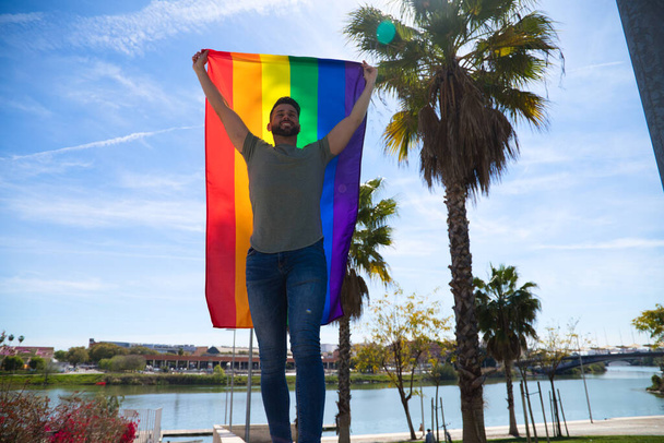 Красивый молодой гей с флагом гей-гордости на ветру, голубое небо на заднем плане. Флаг развевается на ветру. Понятие гомосексуализма, равенства, прав. 28 июня - Фото, изображение