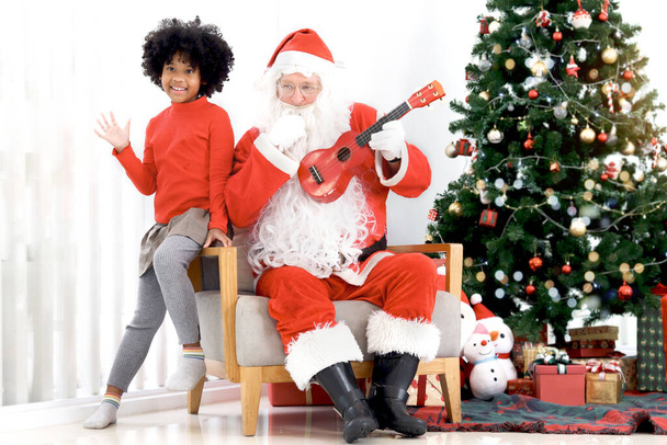 Santa Claus jugando Ukulele con el niño de pelo de curry afroamericano feliz cantando canciones de Navidad frente al árbol de Navidad decorativo en la sala de estar, feliz celebración de vacaciones de invierno. - Foto, imagen