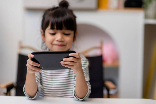 Cute little girl uses smartphone while sitting at the sofa in the living room. Child surfing the internet on mobile phone, browses through internet and watches cartoons online at home - Fotó, kép