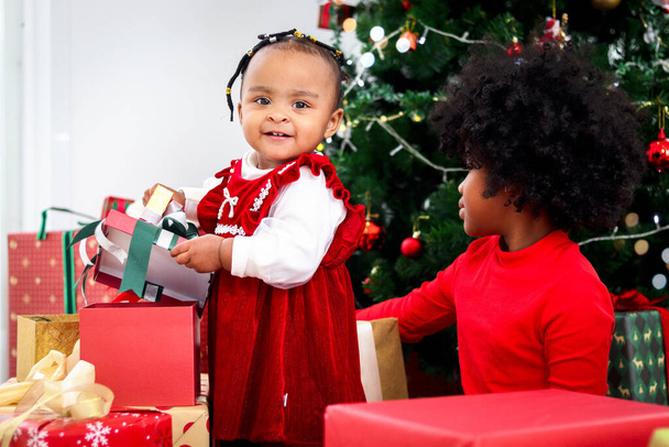 Twee schattige vrolijke glimlachende Afro-Amerikaanse kleine meisje zusjes met zwart curry haar bevatten veel cadeautjes onder kerstboom in de woonkamer, kind vieren vrolijke kerstvakantie - Foto, afbeelding