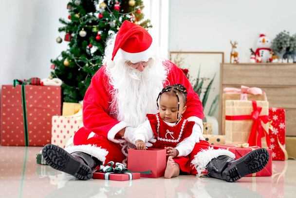 Adorable happy smiling African American child girl sitting on Santa Claus lap around decorative Christmas tree, kid open Christmas gift box present, feeling surprised and excited - Photo, Image