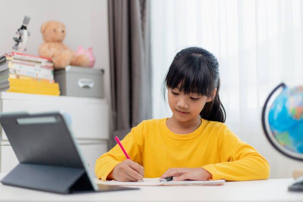 Asyalı liseli kız evde dijital tabletle ödevini yapıyor. Çocuklar çalışmak için aletler kullanırlar. Çocuklar için eğitim ve uzak mesafe eğitimi. Karantina sırasında evde eğitim. Evde kal. - Fotoğraf, Görsel