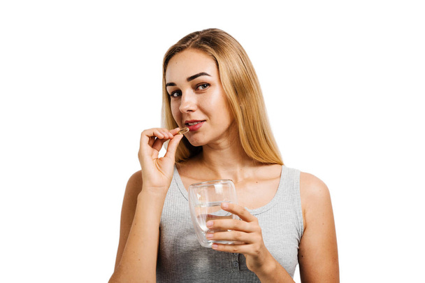 Dívka s kapslí Omega-3 a šálkem vody užívá tobolku BADS na bílém pozadí. Biologicky aktivní doplňky stravy. Užívání vitamínu D pro stavbu a udržování zdravých kostí - Fotografie, Obrázek