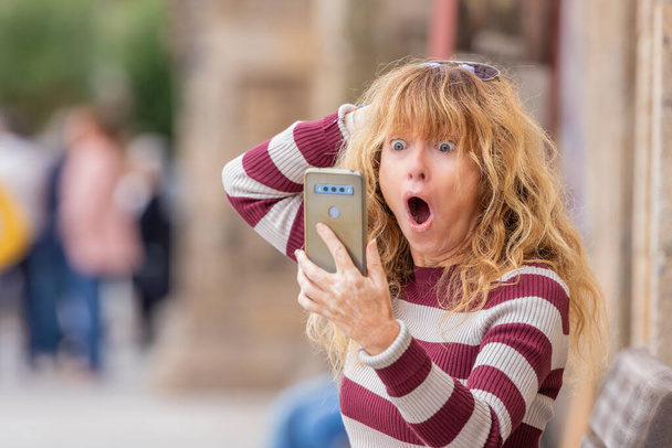 vrouw van middelbare leeftijd met mobiele telefoon in de straat en uitdrukking van verrassing - Foto, afbeelding