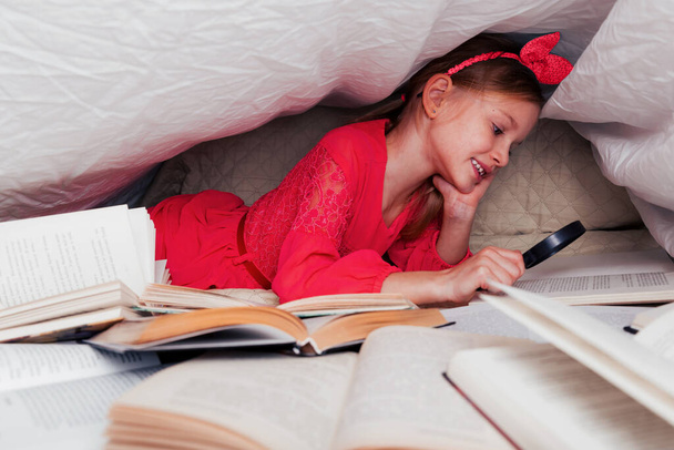 Cute 6-7 years old girl reads a book. Little girl at home on the bed in red dress under a blanket cover with a many books on foreground. Search information in book with magnifying glass - Photo, Image