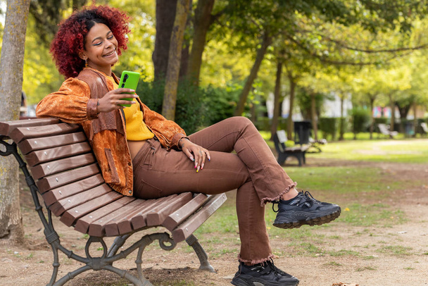 mujer latina negra con teléfono móvil en el banco sentado en otoño - Foto, imagen