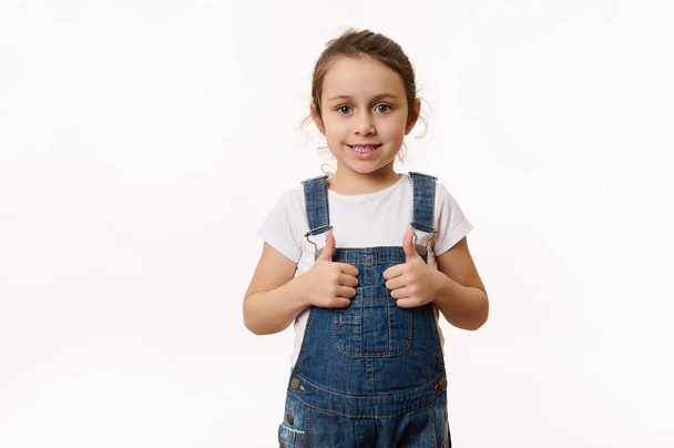 Adorable elementary age Caucasian little child, preschool kid, a lovely baby girl wearing blue denim overalls, shows thumbs up and sweetly smiles looking at camera, isolated on white background - Fotó, kép