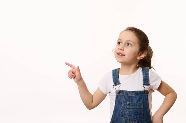 Advertising studio shot of an adorable Caucasian little child, cute baby girl in blue denim overalls, pointing her finger and looking at a copy ad space for promo text, isolated over white background - Photo, Image