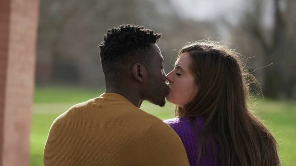 Back of interracial couple kissing sitting at park bench - Фото, изображение