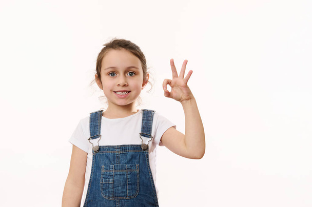 Adorable elementary age, preschool little child girl, in blue denim overalls, gestures, shows peace sign, cutely smiling cheerful toothy smile, looking at camera, isolated white background. Copy space - Photo, Image