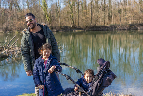 Padre ridente con passeggino e due bambini che passeggiano lungo il fiume Aare nella foresta ripariale in primavera. - Foto, immagini