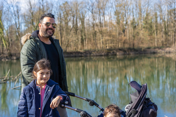 春の阿里川沿いを歩くベビーカーと2人の子供を持つ父親のクローズアップ. - 写真・画像
