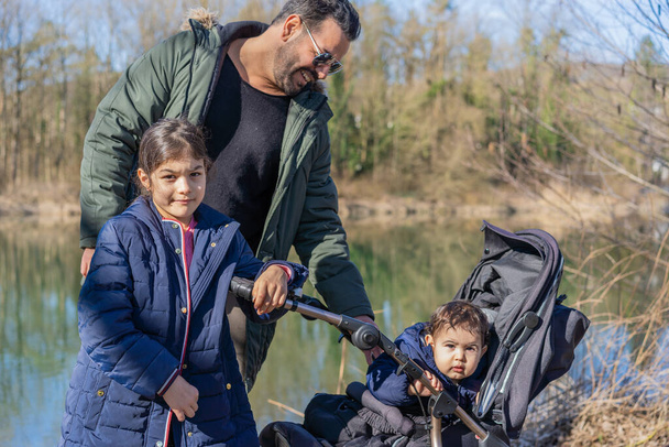 Közelkép apáról babakocsival és két gyerekről, akik az Aare folyó mentén sétálnak tavasszal.. - Fotó, kép