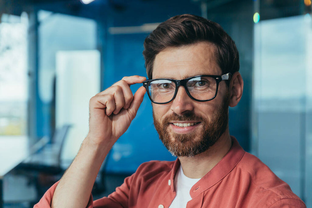 Foto de primer plano del joven arquitecto exitoso con barba, diseñador masculino maduro mirando a la cámara y sonriendo en gafas y camisa roja cerca de la ventana dentro de la oficina. - Foto, Imagen