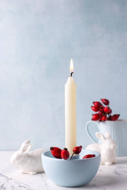 White burning candle, red briar berries  and white bunnies on white marble background against blue  textured wall. Rabbit is symbol of 2023.. Selective focus. Place for text. - Foto, imagen