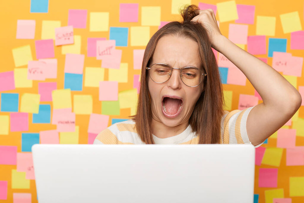 Shocked worried woman with brown hair working on laptop against yellow wall with memo cards, having problems with her online work, lost all information from her computer, pulls her hair, screaming. - Zdjęcie, obraz