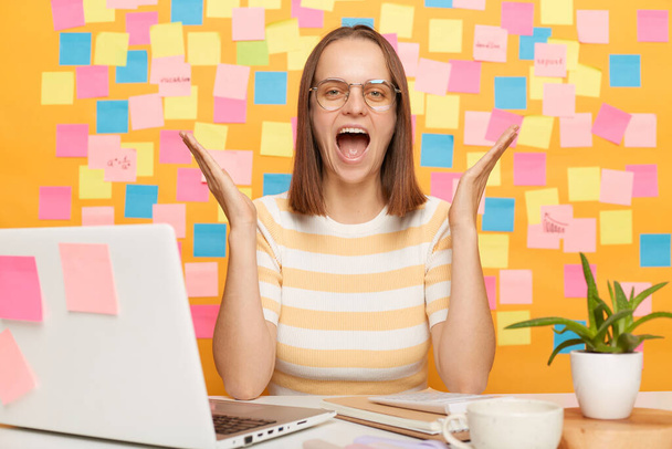 Shocked woman in eyeglasses sitting at workplace in front of notebook, having troubles with internet connection needs work, screaming keeps arms raised, posing against yellow wall with memo cards. - Fotoğraf, Görsel