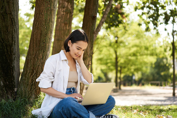 Happy young asian girl sits in park near tree, looking at laptop, working remotely from outdoors, talking to someone, video chat. - Photo, Image