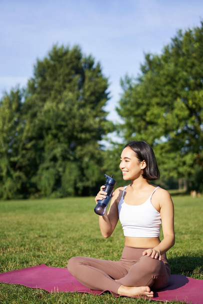 Souriant jeune femme asiatique est assis sur l'herbe, faire des exercices dans le parc, séance d'entraînement sur la pelouse, boit de l'eau de la bouteille. - Photo, image
