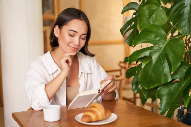 Hymyilevä kaunis tyttö, jolla on kirja, lukee huomaavaisesti virne, ajattelee jotain, juo kahvia kahvilassa.. - Valokuva, kuva