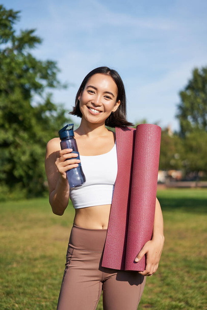 Portrait of young slim and healthy korean girl doing workout in park, standing with water bottle and rubber mat for execises on green lawn, smiling happily. - 写真・画像