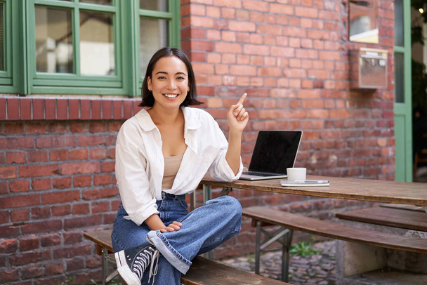 elegante ragazza asiatica moderna con computer portatile, seduto nel caffè, guardando stupito e indicando in alto a destra banner, mostrando pubblicità info. - Foto, immagini