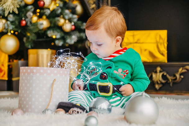 Ein süßer kleiner Junge im Elfenkostüm öffnet ein Weihnachtsgeschenk in einem geschmückten Weihnachtshaus. - Foto, Bild