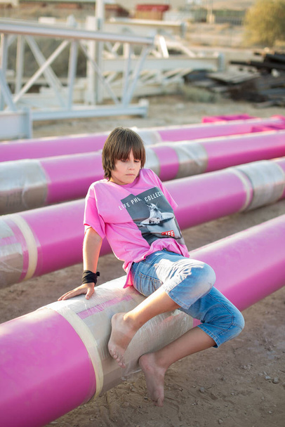 Cute kaukaski chłopiec pozowanie w różowy stylowy drukowany t-shirt wśród rury żelaza różowy na tle przemysłowym. Zdjęcia w stylu mody - Zdjęcie, obraz