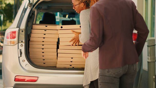 Pracownik Pizzerii rozdaje stosy pudełek po pizzy różnym klientom, wyjmując paczki z bagażnika. Dostawa żywności kurier dostarczanie stosy fast food do klientów czekających w pobliżu drzwi wejściowych. - Zdjęcie, obraz
