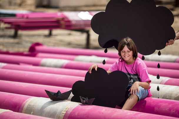 roztomilé dítě v růžovém tričku s kočičím potiskem hraje mezi růžovými trubkami s černými mraky, deštěm, čluny a papírovými letadly proti modré obloze v průmyslovém areálu. focení v módním stylu - Fotografie, Obrázek