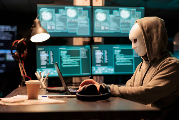 Διαδικτυακός τρομοκράτης με μασκοφόρο χακάρισμα ταυτότητας, που δρα μυστηριωδώς και προσπαθεί να μπει στο σύστημα υπολογιστών. Hacker φορώντας μάσκα και κουκούλα για να κλέψει πληροφορίες online βάση δεδομένων. - Φωτογραφία, εικόνα