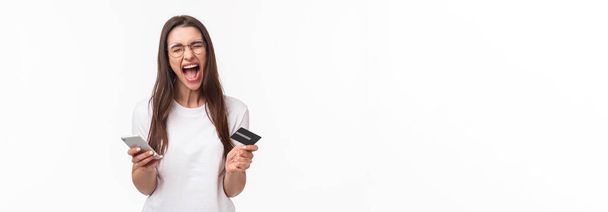 Portrait of uneasy annoyed and angry young brunette female in t-shirt yelling pissed-off as checking bank account, have no money on credit card, or it expired, hold mobile phone, white background. - 写真・画像