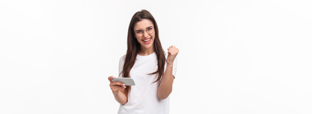 Communication, technology and lifestyle concept. Portrait of successful cheerful lucky girl winning prize in game, finished hard level, fist pump after win on mobile phone app, white background. - 写真・画像