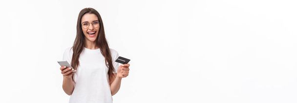 Portrait of happy smiling pretty woman in glasses holding credit card, mobile phone, laughing at camera easily paying for her purchase online store, buy things with application, white background. - Foto, imagen
