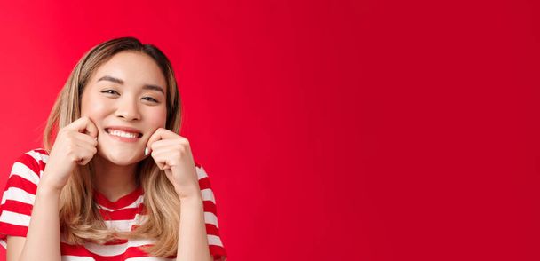 Vidám boldog optimista ázsiai lány, hogy széles örömteli mosoly nyújtózkodik tiszta őszinte vigyor megjelenés kamera elégedett élvezni nyári szünidő squeez arc érintés tiszta tiszta arc, piros háttér. - Fotó, kép