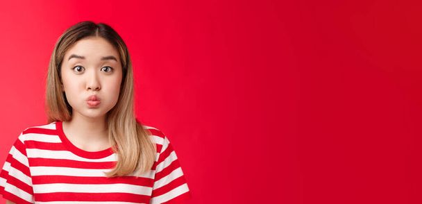 Close-up silly cute asian blond girl pouting making playful face, fold lips hold breath stand red background speechless, stay silent posing childish carefree look impressed. Copy space - Photo, Image