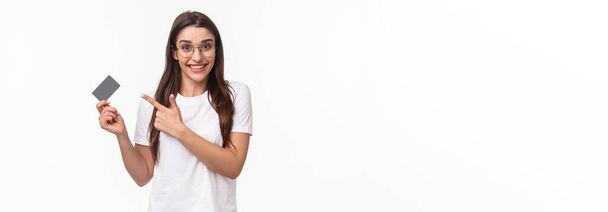 Portrait of excited, happy young woman talking about her bank, pointing at credit card and smiling camera, advice use this for non-cash payment, purchasing goods online, white background. - Photo, Image