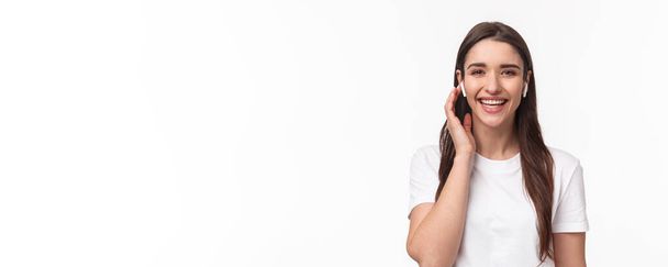 Κοντινό πλάνο πορτρέτο της ελκυστικής χαμογελαστή ευτυχισμένη γυναίκα, αγγίζοντας ασύρματα ακουστικά, χρησιμοποιώντας ακουστικά για να επικοινωνήσετε με το φίλο, έχοντας τηλεφωνική συνομιλία με ακουστικά, γελώντας ανέμελα. - Φωτογραφία, εικόνα