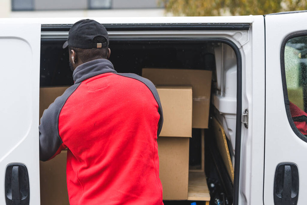 Black young adult delivery man in work uniform unloading cardboard box packages from white van. Horizontal rear view outdoor shot . High quality photo - Фото, зображення