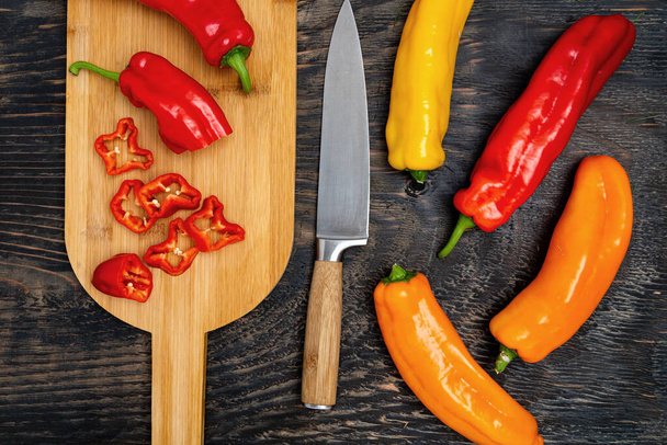 Fresh raw sweet pepper (bell pepper) on a cutting board with a knife. Chopped pepper pieces are ready to cook. Dark wooden table, top view. - Φωτογραφία, εικόνα