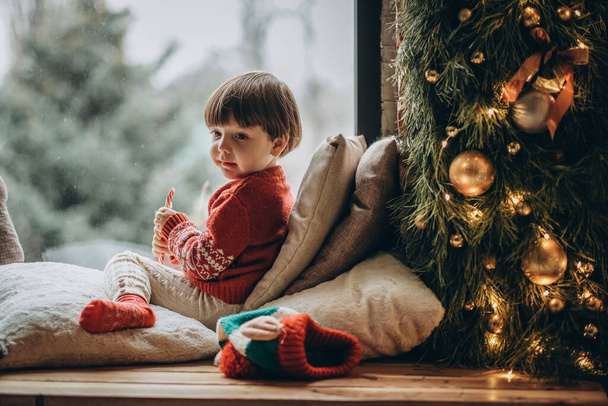 Το παιδί κάθεται στο παράθυρο και περιμένει τον Άγιο Βασίλη. Περιμένω τις γιορτές των Χριστουγέννων. Ευτυχισμένη παιδική ηλικία. Αναμονή για πυροσβέστες. - Φωτογραφία, εικόνα