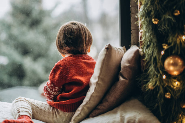Το παιδί κάθεται στο παράθυρο και περιμένει τον Άγιο Βασίλη. Περιμένω τις γιορτές των Χριστουγέννων. Ευτυχισμένη παιδική ηλικία. Αναμονή για πυροσβέστες. - Φωτογραφία, εικόνα